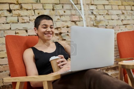 cheerful businesswoman holding coffee to go and using laptop, coworking, startup culture, gen z