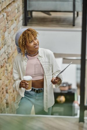 mujer afroamericana feliz con frenos sosteniendo taza de papel, café y documentos, proyecto de puesta en marcha