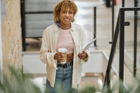 mujer afroamericana positiva con frenos sosteniendo taza de papel, café y documentos, proyecto de inicio
