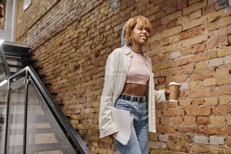 mujer afroamericana feliz con frenos sosteniendo papeles, proyecto de puesta en marcha, caminar en coworking