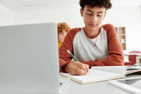 Teenage schoolboy writing on notebook near laptop and blurred classmate in classroom in school