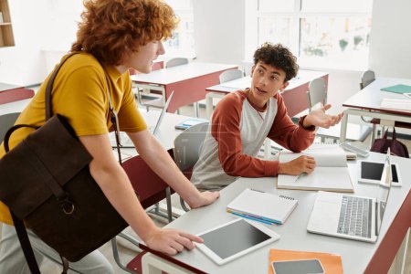 Teenager im Gespräch mit Freund mit Rucksack in der Nähe von Geräten und Notizbüchern auf Schreibtisch im Klassenzimmer