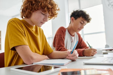 Redhead schoolboy writing on notebook near devices and blurred classmate in school at background