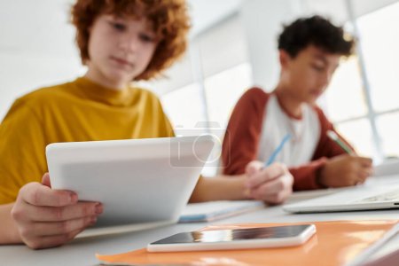 Escolar adolescente borrosa usando tableta digital cerca de amigo y portátil durante la lección en el aula