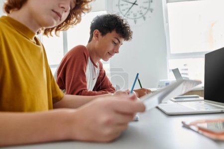 Teenager-Schüler schreibt, während er neben Klassenkamerad sitzt und Geräte im Klassenzimmer im Hintergrund