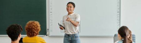 Téléchargez les photos : Professeur afro-américain souriant avec dispositif pointant vers les élèves pendant les cours en classe, bannière - en image libre de droit