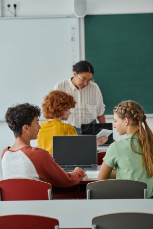 Teen classmates sitting and talking near laptop with blank screen during lesson in class in school