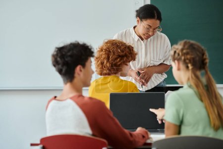 Foto de Profesor afroamericano de pie cerca de alumnos borrosos con computadoras portátiles durante la lección en el aula - Imagen libre de derechos