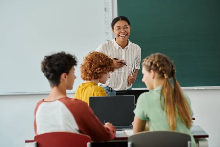 Profesor afroamericano sosteniendo cuaderno cerca de los alumnos con dispositivos durante la clase en la escuela