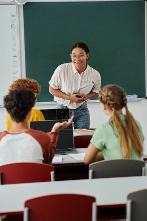 Professeur afro-américain souriant avec cahier regardant un écolier pendant les cours en classe