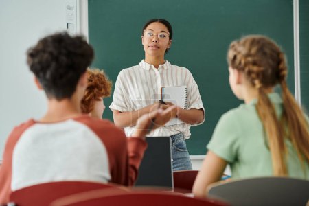 Pensativo profesor afroamericano sosteniendo cuaderno cerca de los alumnos y el ordenador portátil en el aula