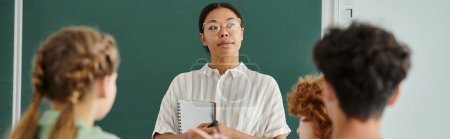 Pensive african american teacher holding notebook near blurred pupils in classroom, banner