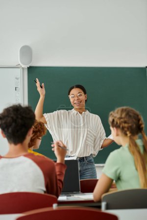 Profesora afroamericana positiva hablando con alumnos adolescentes borrosos cerca de la computadora portátil en el aula en la escuela