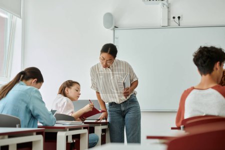 Téléchargez les photos : Écolière adolescente montrant un cahier à un professeur afro-américain pendant un cours en classe à l'école - en image libre de droit