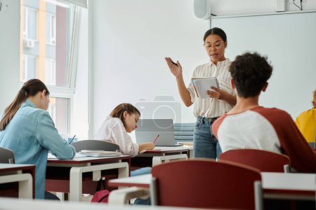 Téléchargez les photos : Professeur afro-américain avec ordinateur portable pointant et parlant pendant les cours avec des élèves adolescents en classe - en image libre de droit