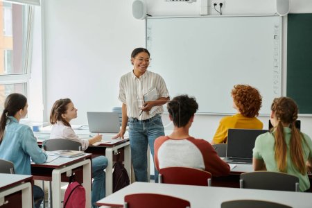 Téléchargez les photos : Professeur afro-américain positif avec un cahier debout près des élèves et des appareils en classe - en image libre de droit