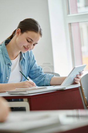Alumno adolescente sonriente usando tableta digital y escribiendo en un cuaderno durante la lección en el aula