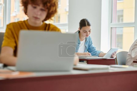 Teenager-Schülerin hält digitales Tablet in der Hand und schreibt auf Notizbuch in der Nähe verschwommener Mitschülerin im Klassenzimmer