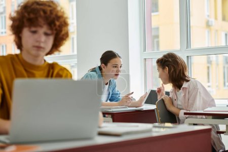 Foto de Compañeros de clase adolescentes hablando y señalando con los dedos cerca de dispositivos y cuaderno en el aula - Imagen libre de derechos