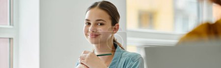Cheerful teenage schoolgirl holding pen and looking at camera during lesson in class, banner