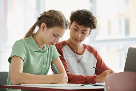 Teen schoolboy writing and looking at notebook near classmate during lesson in classroom in school