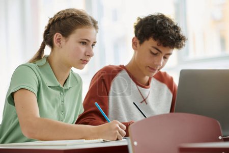 Teenager-Schülerin schreibt während des Unterrichts auf Notizbuch neben verschwommenem Klassenkameraden und Laptop