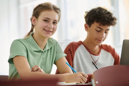 Smiling teen schoolgirl looking at camera while writing on notebook near laptop and friend in class