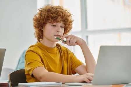 Redhead teenage schoolboy holding pencil and using laptop during lesson in classroom at background