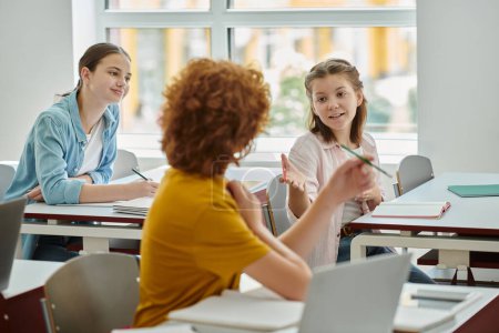 Positive teen schoolgirl pointing with hand while talking to classmate near devices in classroom