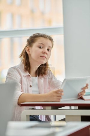 Focused teenage schoolgirl holding digital tablet and looking at camera during lesson in classroom