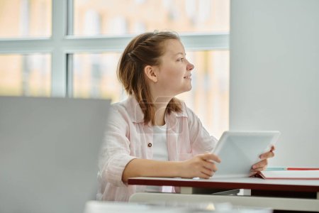 Photo for Side view of smiling teen schoolgirl holding digital tablet and looking away during lesson in class - Royalty Free Image