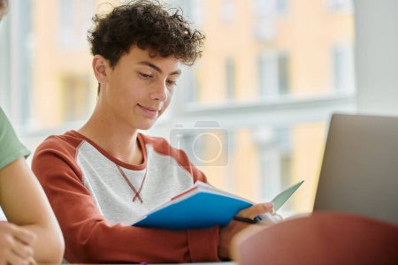 Curly teenage schoolboy looking at notebook near classmate and laptop in classroom in school