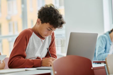 Photo for Curly teenage schoolboy writing on notebook near laptop during lesson in classroom in school - Royalty Free Image