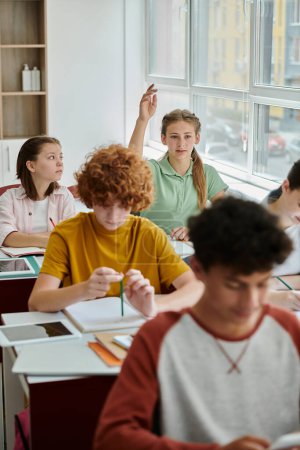 Adolescente escolar levantando la mano y hablando cerca de dispositivos y compañeros de clase durante la lección en la escuela