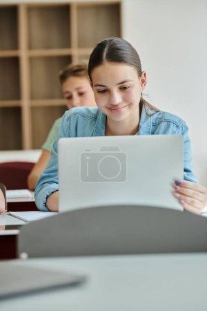 Adolescente positiva colegiala en ropa casual usando el ordenador portátil durante la lección en clase en la escuela