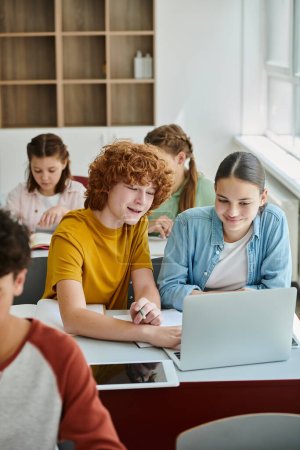Foto de Sonriendo compañeros de clase adolescentes que utilizan el ordenador portátil juntos cerca de cuadernos durante la lección en el aula en la escuela - Imagen libre de derechos