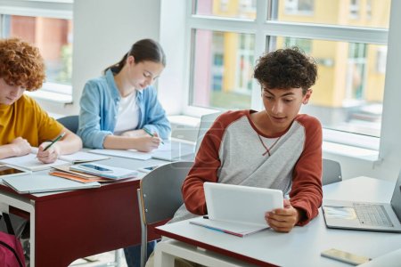 Teenager mit digitalem Tablet in der Nähe von Geräten mit leerem Bildschirm und Mitschülern im Klassenzimmer
