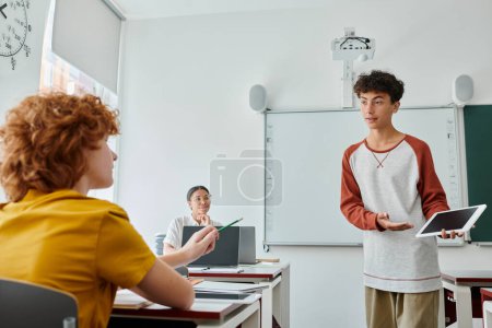 Teen schoolboy pointing at digital tablet with blank screen near classmate during lesson in class