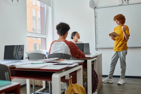 Redhead teen schoolboy holding book and talking near african american teacher during lesson in class