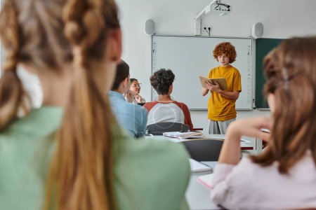 Redhead pupil holding book and talking near african american teacher and friends in classroom