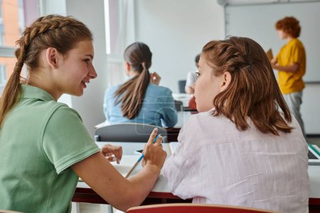 Lächelnder Teenager im Gespräch mit Klassenkamerad und Zeigefinger während des Unterrichts im Klassenzimmer
