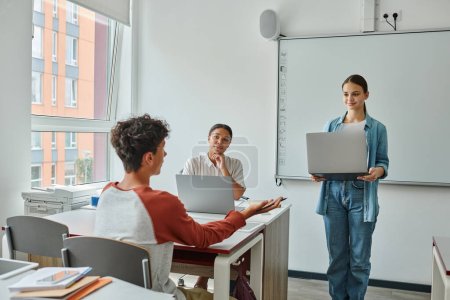Teen schoolboy talking to classate with laptop near african american teacher in classroom
