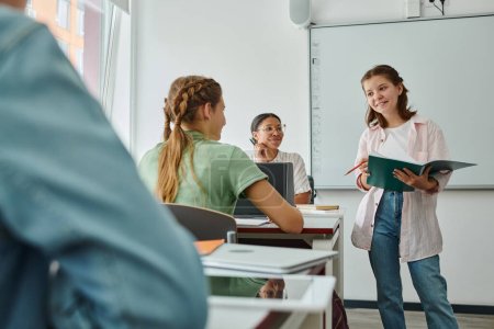 Photo for Positive teen schoolgirl holding notebook near classmate and african american teacher in classroom - Royalty Free Image