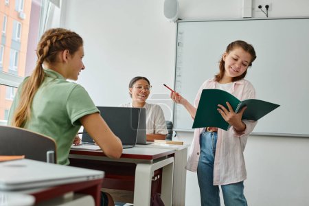 Smiling schoolgirl talking and holding notebook near african american teacher and classmate in class