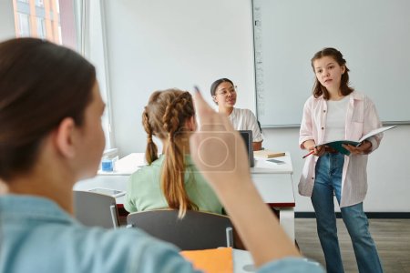 Teen schoolgirl holding notebook near classmate raising hand and african american teacher in class