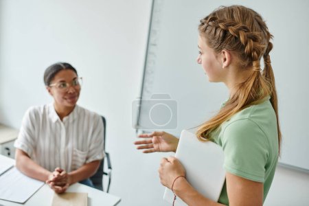 Photo for Teen schoolgirl holding digital tablet and talking to blurred african american teacher in class - Royalty Free Image