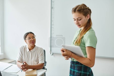 Adolescente colegiala usando tableta digital cerca borrosa africano americano profesor durante lección