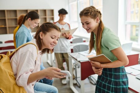 Escolar adolescente sonriente con mochila mostrando smartphone a amigo con portátil en el aula