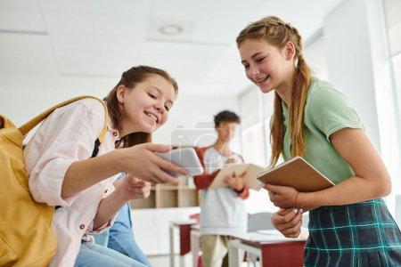 Smiling teen schoolgirls with backpack and notebook using smartphone in classroom in school
