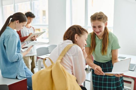 Téléchargez les photos : Joyeux écolière tenant cahier proche ami avec smartphone et sac à dos dans la salle de classe - en image libre de droit
